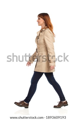 Confident young woman is walking in beige coat, jeans and brown shoes. Side view. Full length studio shot isolated on white.