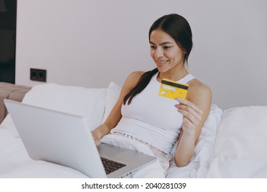 Confident Young Woman In White Tank Top Sit In Bed Using Laptop Pc Computer Holding Credit Bank Card Doing Online Shopping In Bedroom Lounge Home In Own Room House Wake Up Be Lost In Reverie Good Day