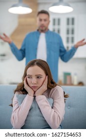 Conflict. Dad Raising His Hands To The Side Standing Behind The Sad Daughter Sitting On The Couch, Resting His Face In The Palm Of His Hands.