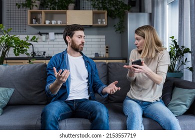 Conflict, quarrel, jealousy in the family home. Young couple, man and woman. The woman is jealous, takes the phone from her husband, demands an explanation - Shutterstock ID 2153557719