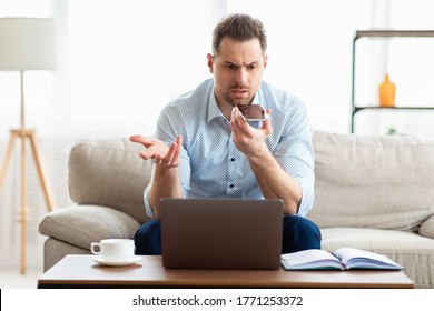 Confused adult businessman talking on phone, holding cellphone near mouth and gesturing, looking at laptop