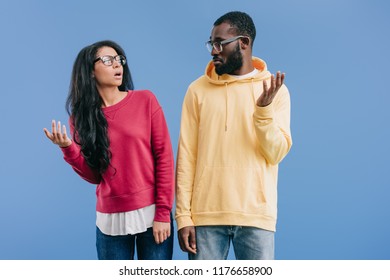 Confused African American Couple In Eyeglasses Looking At Each Other And Doing Dubium Gesture Isolated On Blue Background 