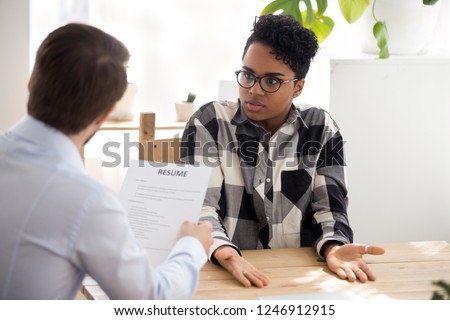 Confused African American millennial female applicant shrug shoulder have misunderstanding with employer or HR manager, businessman consider black candidate resume accuse or blame her for mistake
