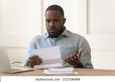 Confused African Man Sit At Desk Hold Mail Paper Received Letter Read Shocking Unexpected News, Guy Feels Mad About High Bill Tax Invoice, Debt Notification From Bank, Bad Financial Situation Concept