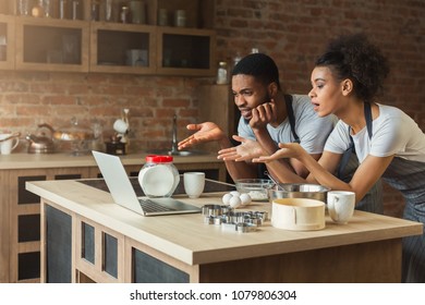 Confused African-american Couple Cooking Dinner With Recipe, Using Laptop In Their Loft Kitchen At Home. Preparing Some Pasrty