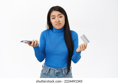 Confused Asian Girl Holds Smartphone With Credit Card, Shrugs Shoulder With Puzzled Face, White Background.