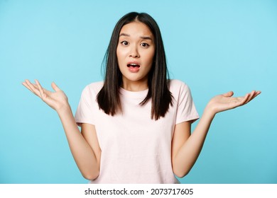 Confused asian girl spread empty hands sideways, shrugging shoulders clueless, standing in tshirt over blue background - Shutterstock ID 2073717605
