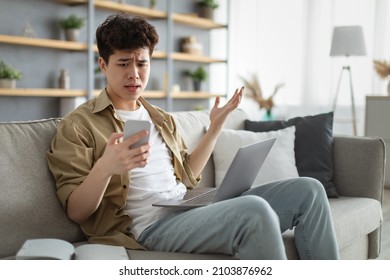 Confused Asian Man Looking At Phone Screen, Sitting On Couch With Computer On Lap In Living Room, Upset Frustrated Male Reading Bad News On Message, Having Problem With Device Or Internet Connection