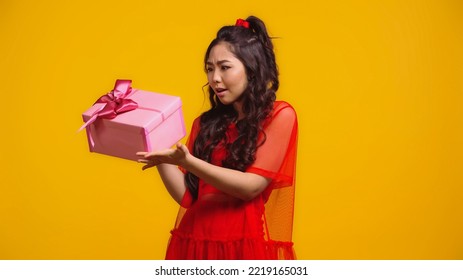 Confused Asian Woman Holding Wrapped Present Isolated On Yellow 