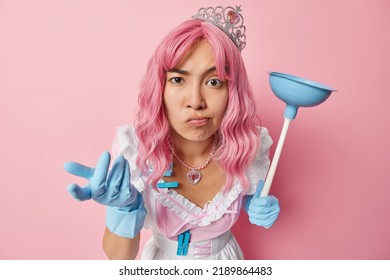 Confused Asian Woman With Long Dyed Hair Holds Plunger Looks Unaware Shrugs Shoulders Wears Crown Dress And Rubber Gloves Does Housework Isolated Over Pink Studio Background. So What To Do Now