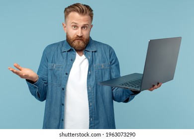 Confused bearded blonde hipster young man in jeans shirt holding open laptop on blue background.