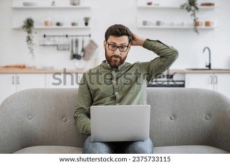Confused bearded male wearing eyeglasses and casual attire shrugging shoulders while sitting on couch with modern laptop and looking at camera. Concept of doubt, people and technology.