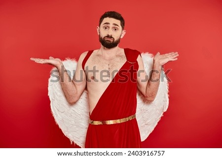 confused bearded man in cupid costume showing shrug gesture and looking at camera on red backdrop