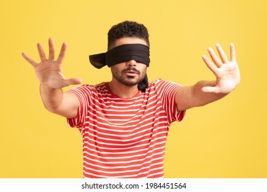 Confused bearded man in red striped t-shirt with blindfold on eyes moving in darkness with outstretched arms, training, developing senses. Indoor studio shot isolated on yellow background
