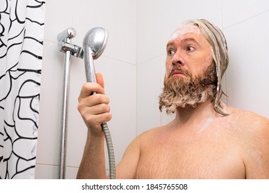 Confused bearded man with soapy head standing in the bathroom and looking at the shower while the water supply has stopped. No water coming from tap