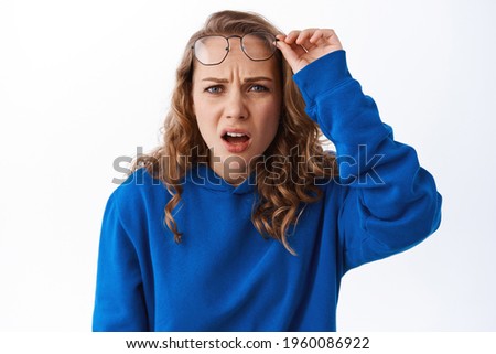 Confused blond girl take off glasses, look with puzzled and displeased face, cringe from something strange and awful, standing in blue hoodie against white background