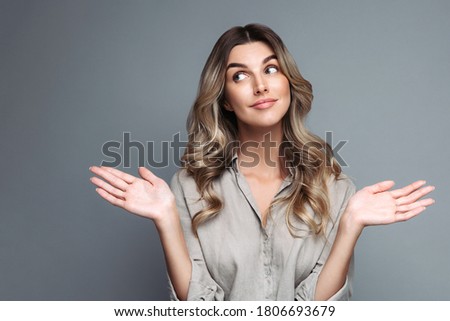 Confused blonde woman with curly hair dressed in linen shirt spreads hands sideways and shrug shoulders, smirks face, looking up at blank space for advertisement, isolated on studio grey background