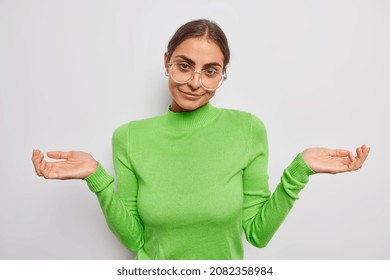 Confused Brunette Young Woman Spreads Palms With Hesitation Has No Clue Or Idea Stands Puzzled Wears Transparent Spectacles Casual Green Turtleneck Isolated Over White Background. So Who Cares