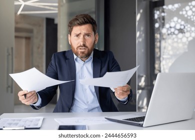 Confused businessman in a suit looks angrily at paperwork in a modern office setting, feeling frustrated - Powered by Shutterstock