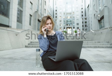 Confused businesswoman talking via smartphone looking at her laptop on the stairs near the office. Puzzled office worker dealing with problems on her computer sitting on the stairs of business center.