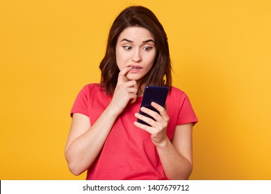 Confused Busy Interested Young Lady Holding Smart Phone In One Hand, Looking At Its Screen Attentively, Putting Forefinger In Mouth, Looks Worried, Posing Isolated Over Yellow Background In Studio.