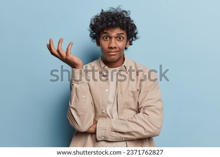 Confused careless man shrugs shoulder with hesitation wears beige shirt says I dont know what to looks clueless poses against blue background. Unsure Hindu guy asks who knows. Difficult choice