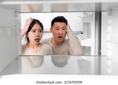 Confused Chinese Couple Looking Inside Open Empty Fridge In Kitchen At Home, Touching Their Heads. Poor Asian Family Have Nothing To Eat, Copy Space. Financial Hangover While Crisis, Poverty Concept