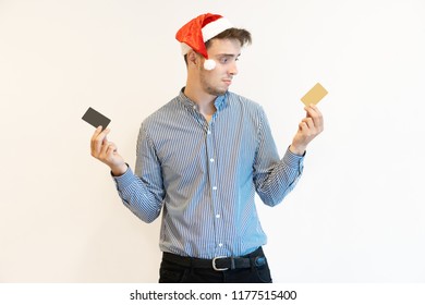 Confused Christmas Cardholder Choosing Between Two Cards. Handsome Young Man In Santa Hat Holding Plastic Cards, One Per Hand. Payment Or Dilemma Concept