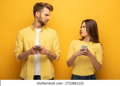 Confused Couple With Smartphones Looking At Each Other On Yellow Background