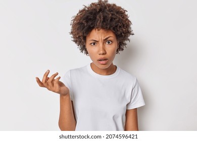 Confused curly haired woman shrugs shoulders cannot understand why says so what being confused and pissed off wears casual t shirt isolated over white background. Human reactions and perception
