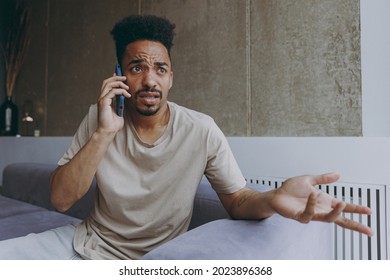 Confused Disappointed Sad Young African American Man 20s In Beige T-shirt Sit On Grey Sofa Indoors Apartment Talk By Mobile Cell Phone Spread Hand Rest On Weekends Stay At Home. Tattoo Translate Fun