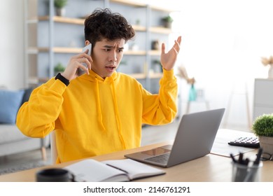 Confused Discontented Asian Guy Talking On Cell Phone Using Pc Having Problem With Computer Or Internet Connection At Home. Displeased Male Customer Calling To Hotline Service And Complaining