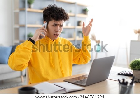 Confused Discontented Asian Guy Talking On Cell Phone Using Pc Having Problem With Computer Or Internet Connection At Home. Displeased Male Customer Calling To Hotline Service And Complaining