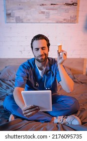 Confused Doctor Holding Pills And Digital Tablet While Suffering From Sleepwalking On Bed