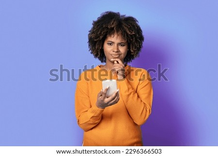 Confused doubtful pretty curly young black woman wearing casual outfit with smatphone in her hand, looking at camera and touching her chin, isolated on purple studio background, copy space