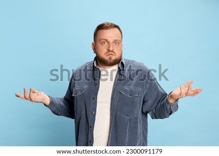 Confused, failure. Nice-looking man wearing jeans jacket spreading hands and looking at camera with face without understanding over blue background. Concept of emotions, facial expression, mood