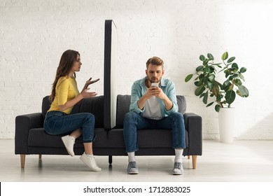 Confused girl looking at big model of smartphone near addicted boyfriend chatting on sofa - Shutterstock ID 1712883025