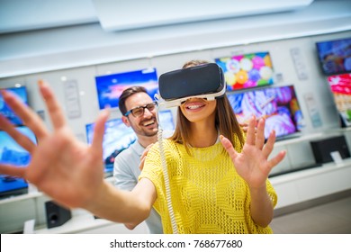 Confused happy young handsome woman tasting VR with hands in front while her attractive smiling boyfriend standing behind her and holding shoulders with hands in a tech store.
