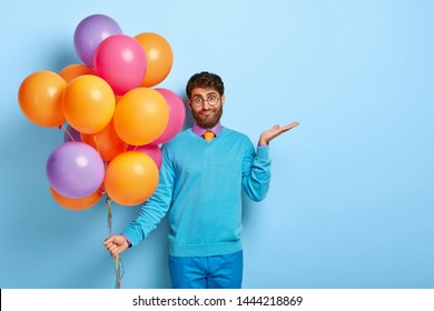 Confused Hesitant Male Holiday Maker Raises Palm With Puzzlement, Tries To Make Decision, Holds Multicolored Bunch Of Air Balloons, Doesnt Know Whether All Guests Has Come, Wears Blue Clothes