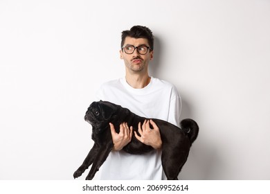 Confused Hipster Guy Holding Black Cute Pug And Looking Left, Feeling Puzzled Or Clueless. Dog Owner Pondering, Carry Dog In Arms, White Background