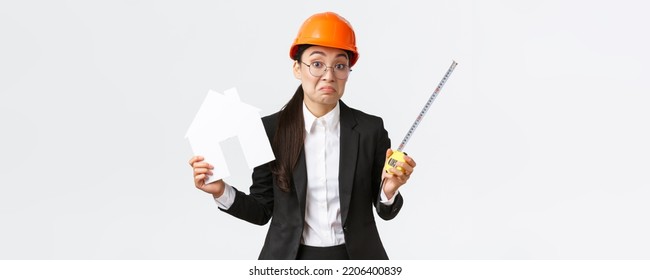 Confused indecisive asian female architect dont know what do, engineer in helmet and business suit shrugging while holding home maket and measuring tape, white background - Powered by Shutterstock