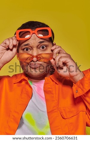 confused indian man trying on different trendy sunglasses and looking at camera on yellow backdrop