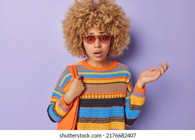 Confused indignant woman with curly hair shrugs shoulders stares shocked at camera wears knitted striped jumper sunglasses carries bag on shoulder isolated over purple background feels puzzled