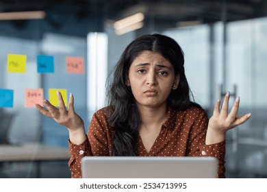 Confused Latina businesswoman shrugs baffled at desk, surrounded by colorful sticky notes. Expression conveys frustration in corporate environment. Represents stress and decision-making challenges. - Powered by Shutterstock