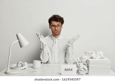 Confused male worker wears everything white, spreads palms, sits at desktop, works with papers, looks with puzzled expression, contemplates about important idea. Executive manager at working place