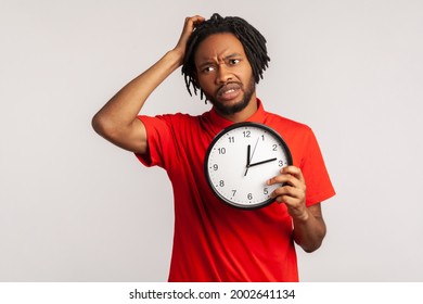 Confused man with dreadlocks wearing red T-shirt, rubbing his head holding in hands big wallclock, has no time , worried about deadline, looking away. Indoor studio shot isolated on gray background.