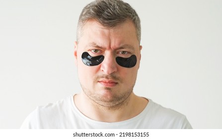 Confused Man. Face Treatment. Anti-aging Procedure. Dissatisfied Gloomy Guy With Hydrogel Black Under Eye Patches On Light Background.