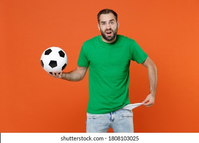 Confused Man Football Fan In Green T-shirt Cheer Up Support Favorite Team With Soccer Ball Has No Money Showing Empty Pocket Has No Money Isolated On Orange Background. People Sport Leisure Concept