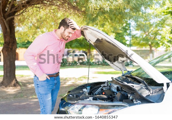 Confused man looking at engine of broken down\
car at roadside