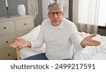 Confused middle-aged man in white shirt shrugging while sitting on a bed in a well-lit bedroom interior.